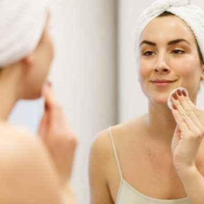 istockphoto-1280518908-612×612 Smiling woman with towel on head after shower looking at her own reflection in the mirror and cleaning face with cotton pad