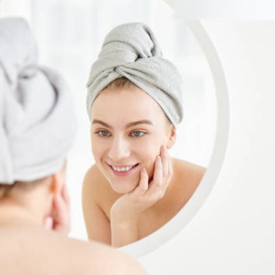 istockphoto-1280707516-612×612 Portrait of young girl with towel on head in white bathroom looks and touches her face in the mirror and enjoys youth and hydration. Natural beauty, home care for problem skin